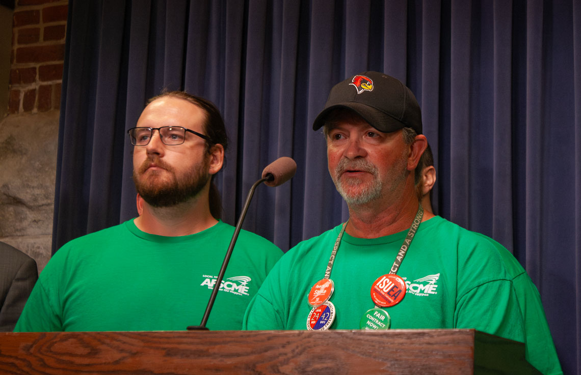 Two men in green speaking at a podium.
