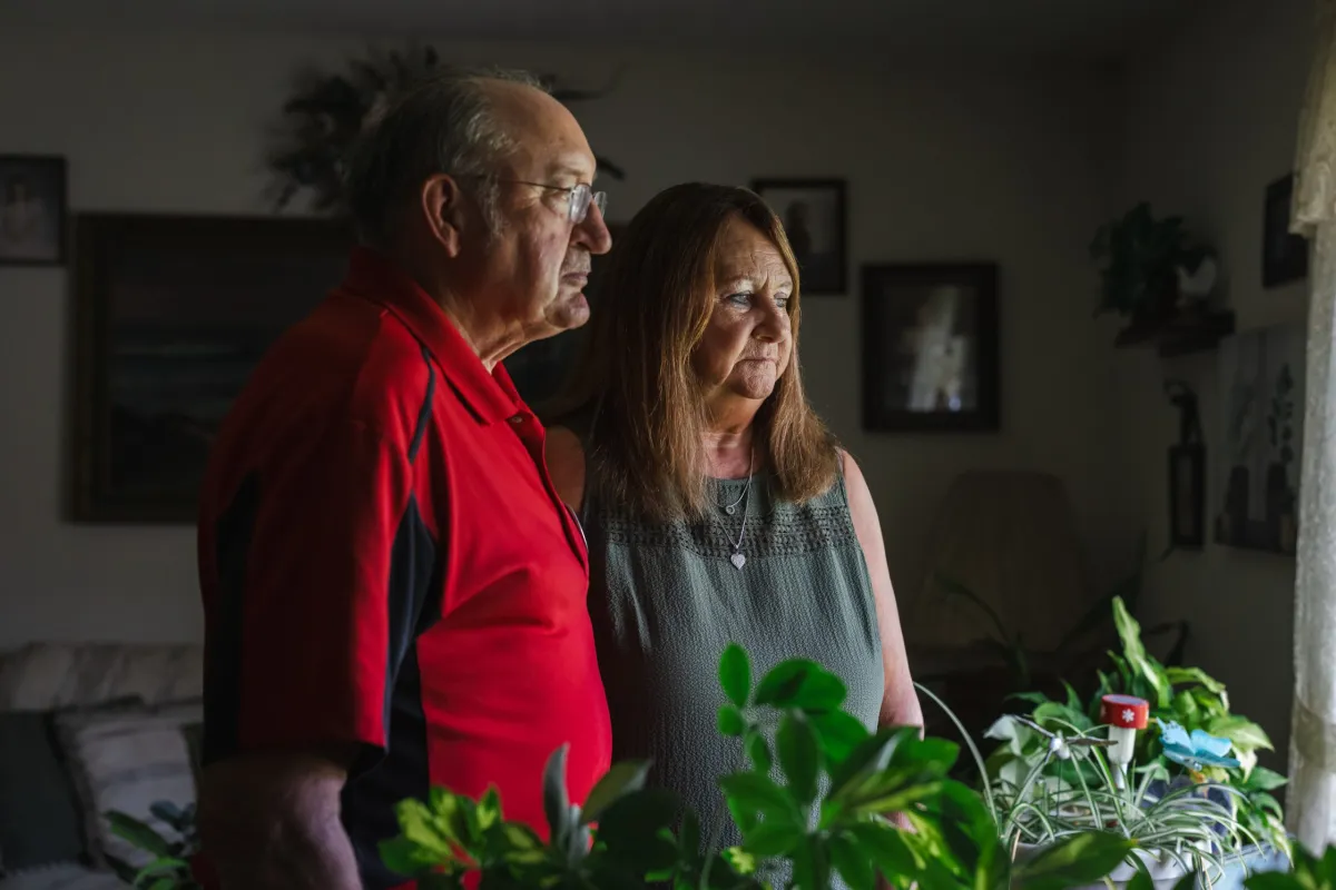 An older couple stands in a dimly lit room, looking off camera past houseplants.